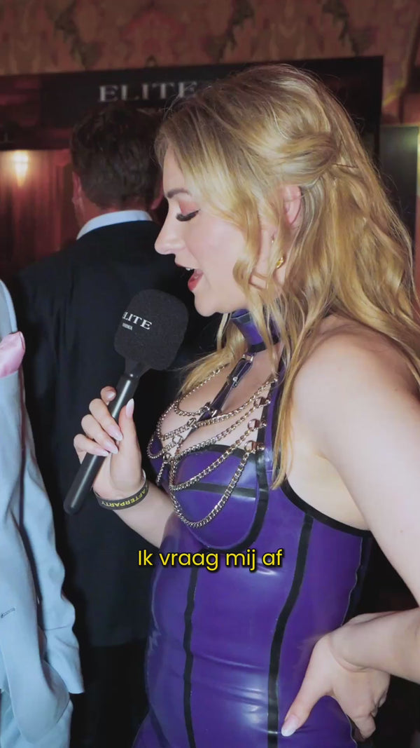 A woman in a striking purple outfit with gold chains holds a black microphone labeled “ELITE” while speaking to someone off-camera. She stands confidently in a warmly lit venue, surrounded by people in formal attire.
